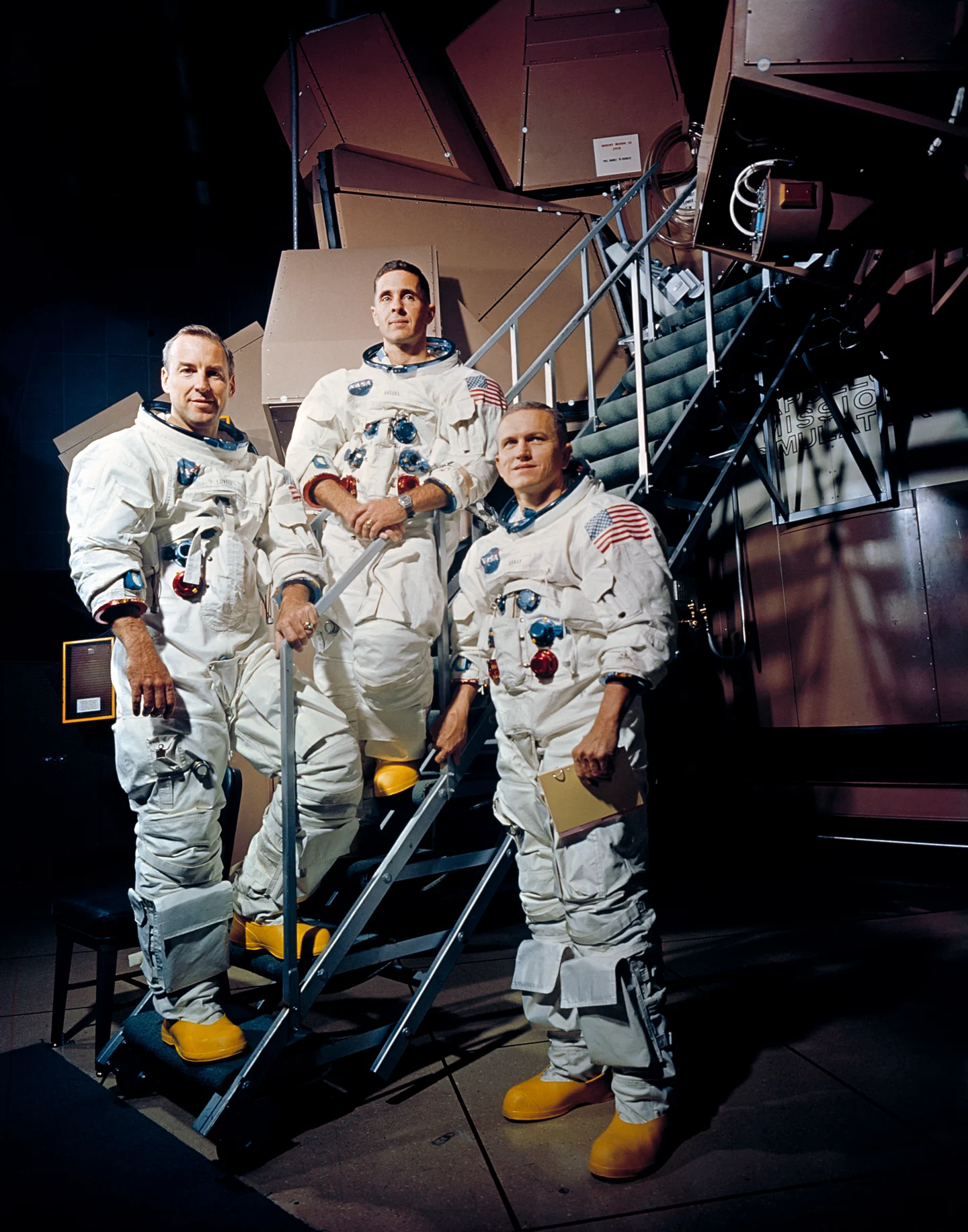 Jim Lovell, Bill Anders and Frank Borman pose in spacesuits beside an Apollo simulator.