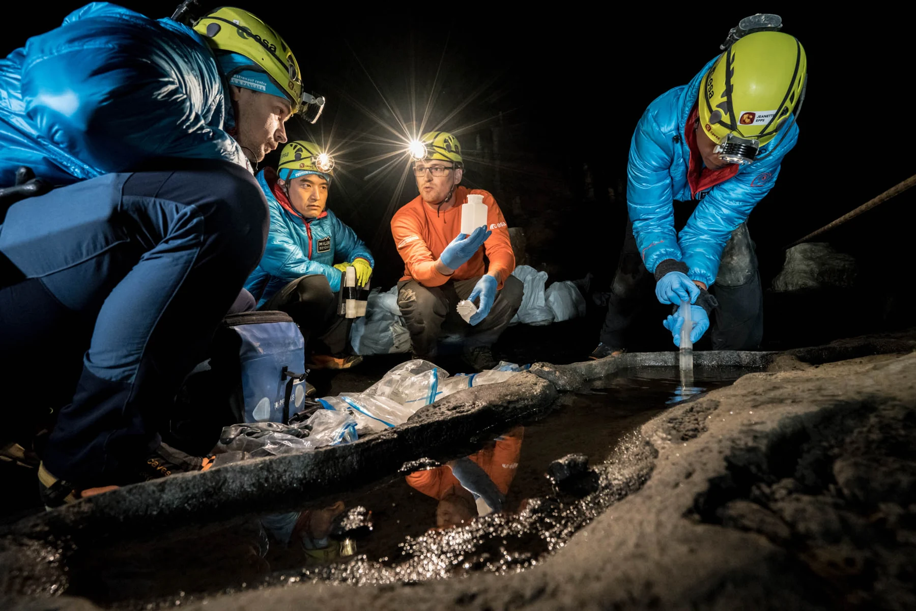 Helmeted CAVES participants gather around a water source while one uses sampling equipment.