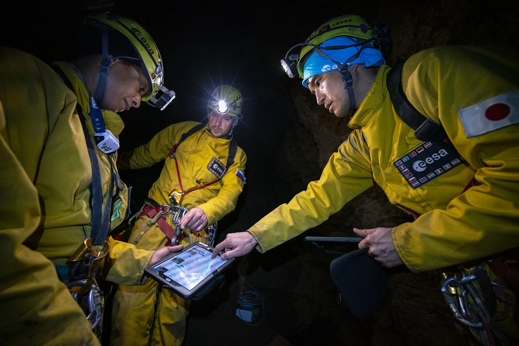 Three helmeted CAVES participants examine a shared tablet in a dark cave.