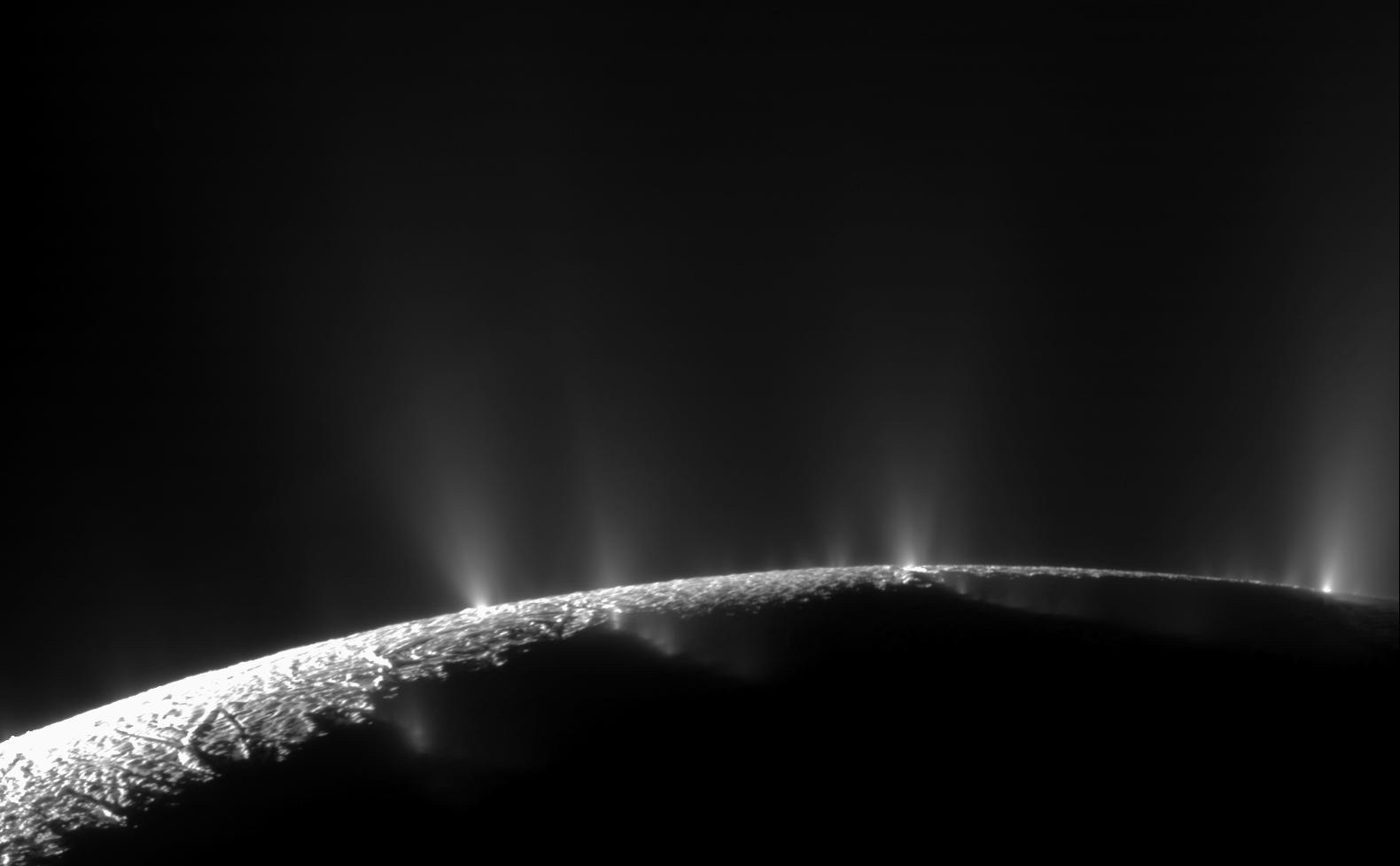 Bright jets rise above the curved, fractured limb of Enceladus in a monochrome mosaic.