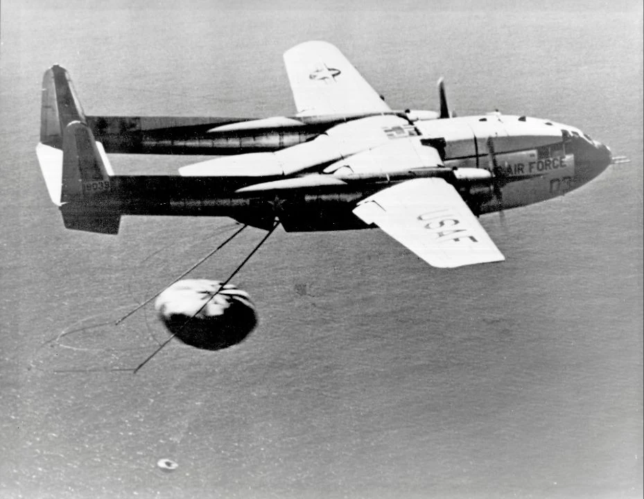 C-119 aircraft above the ocean with recovery poles and a parachute trailing behind its open rear.