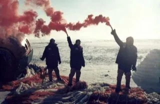 Astronauts hold smoking signal flares beside equipment in a snowy landscape.