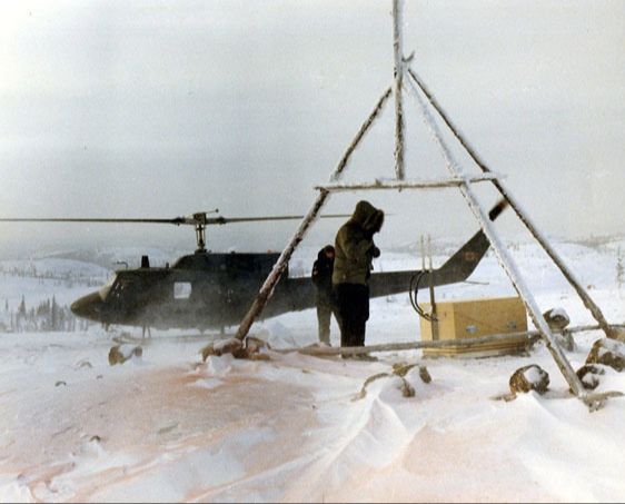 A worker stands under a triangular wooden landmark beside equipment, with a helicopter behind in the snow.