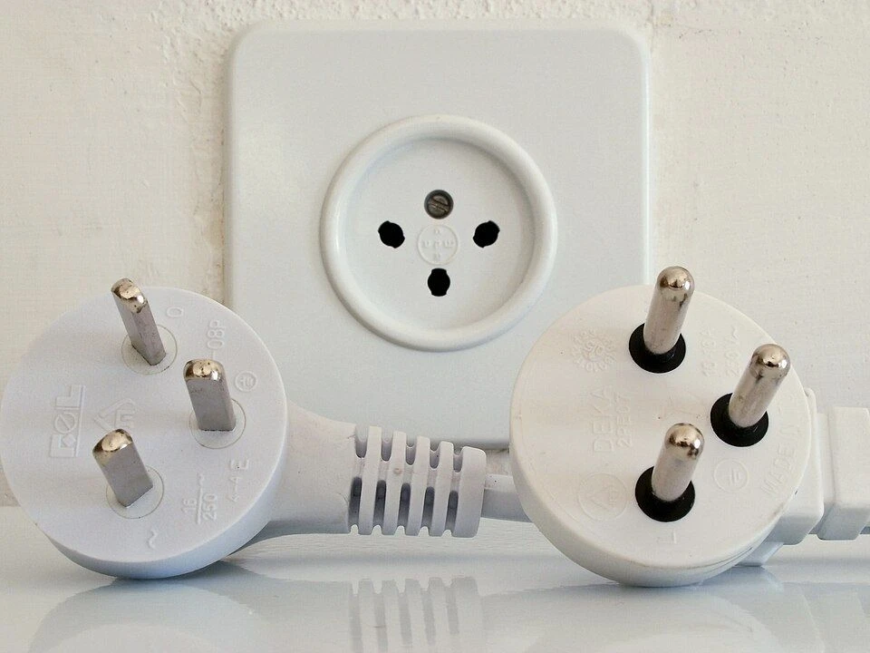 Older flat-pin and newer round-pin Israeli plugs in front of a matching wall outlet.
