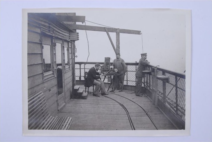 Three people stand and sit beside apparatus on a platform high on the Eiffel Tower.