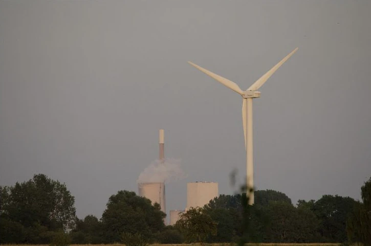 A wind turbine stands in front of a coal-fired power station beyond trees.