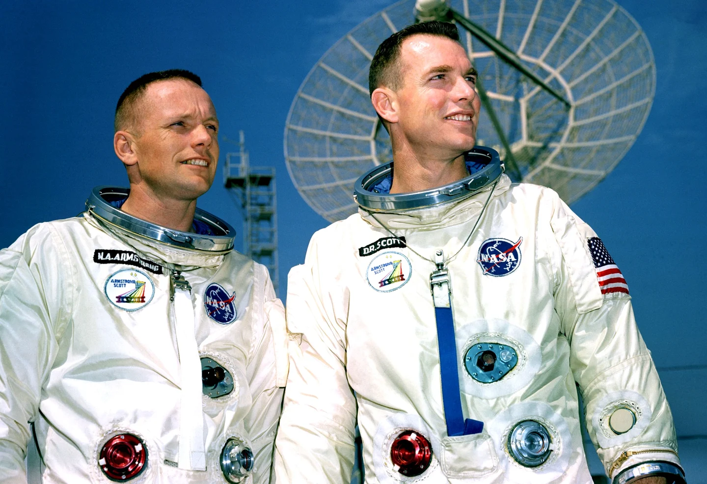 Neil Armstrong on the left and David Scott on the right in spacesuits, standing in front of a radar antenna.