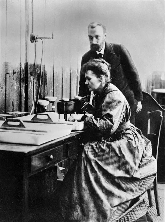 Marie Curie sits at a laboratory bench with measuring instruments while Pierre Curie stands behind her.
