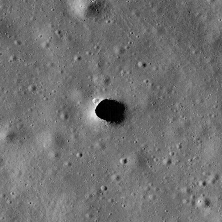 Orbital view of the dark Marius Hills pit amid smaller craters and textured lunar terrain.