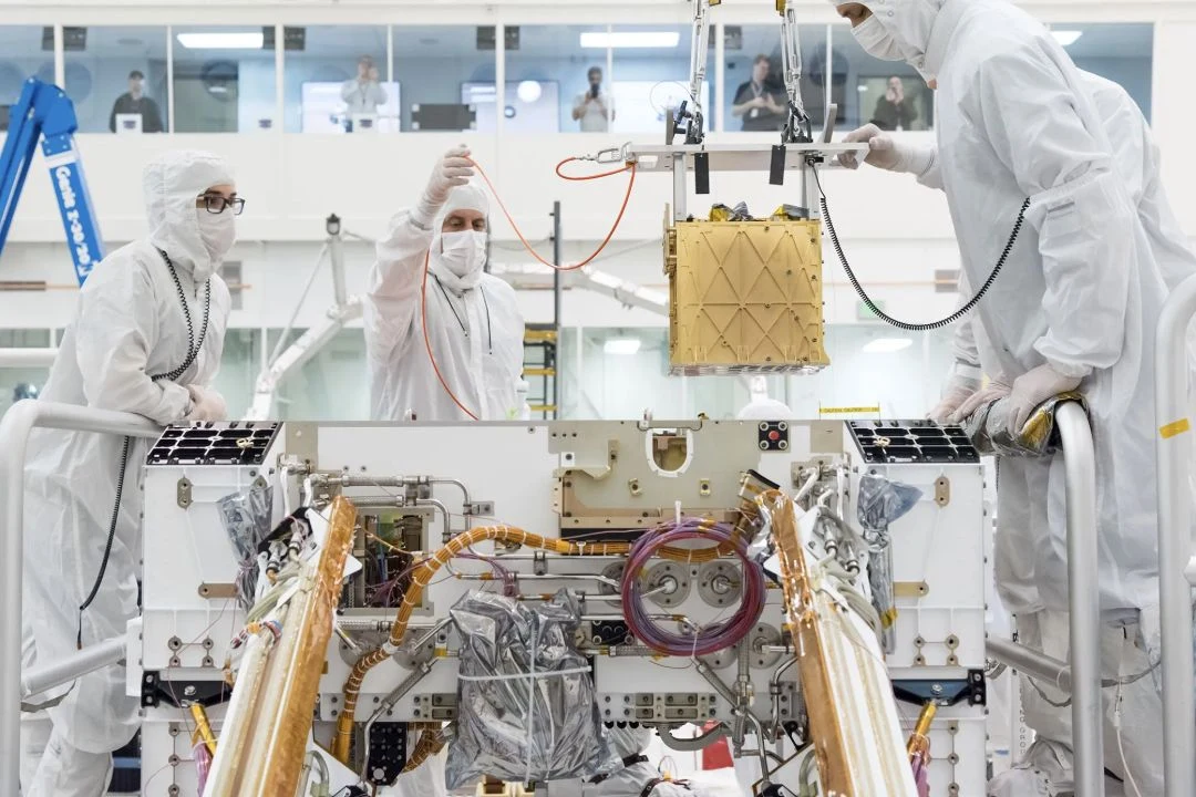 Technicians lower the gold-colored MOXIE instrument into the Perseverance rover in a cleanroom.
