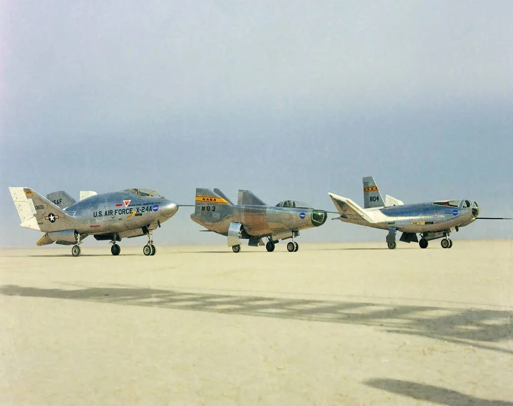Three compact research aircraft stand side by side on a dry lakebed, with broad bodies, fins and wheels but no conventional large wings.