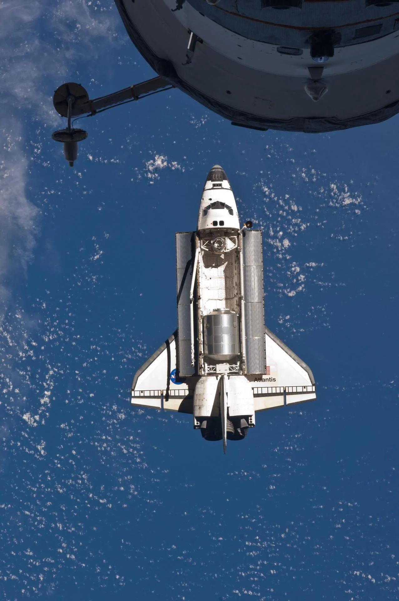 Atlantis approaches the space station with its payload bay open and its broad wings visible; part of a docked Progress vehicle fills the foreground.