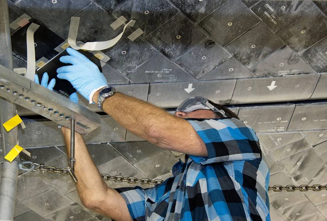 A technician reaches into an opening among the black thermal-protection tiles on Atlantis’s underside.